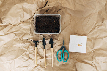 Naklejka premium Kraft paper background, fertile soil and garden tools on top, top view. Planting seedlings in spring