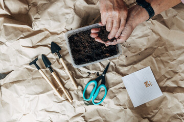 Kraft paper background, fertile soil and garden tools, top view. Hands planting seeds