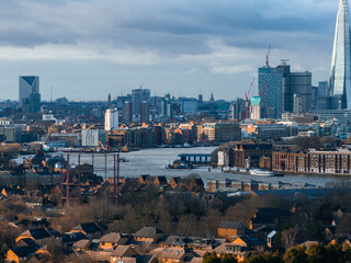 Aerial view of London with the River Thames, the Shard skyscraper, and Canary Wharf. Residential and industrial buildings under a partly cloudy sky.
