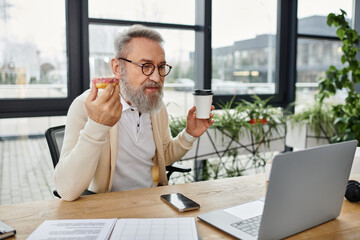 Mature man with a beard holds a pastry and coffee while focusing on his laptop in a sunny office.