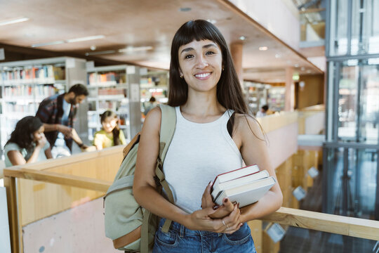 Portrait of smiling woman holding books while standing with bag in university library
