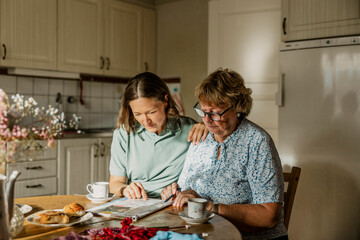 Female nurse helping senior woman in solving crossword puzzle while sitting at dining table
