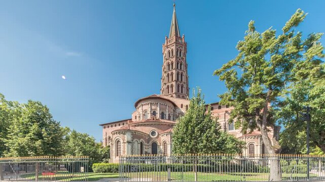 The cobbled parvis of the Basilica of Saint Sernin in Toulouse, France, timelapse hyperlapse. Largest romanesque church in Europe with brick bell tower, arched doors, green trees and blue sky