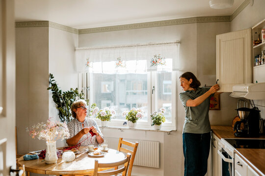 Female caregiver talking with senior woman while opening cabinet in kitchen at home