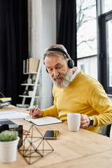 A mature handsome man in a yellow sweater sits at a table, writing notes while sipping coffee.