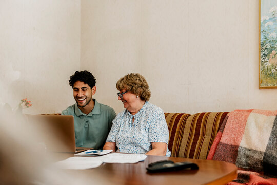 Smiling male nurse using laptop while sitting with senior woman on sofa at home
