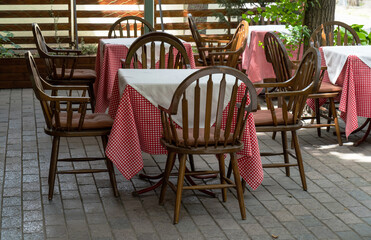 Outdoor summer cafe. Empty tables covered with a checkered white and red tablecloth and wooden chairs on the terrace in front of the cafe, opening onto the street, shaded by a tree. Cobblestone floor.