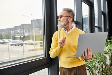 Handsome senior man stands by window holding laptop while looking thoughtfully outside. © LIGHTFIELD STUDIOS