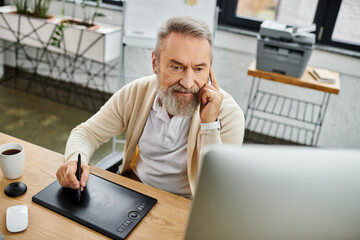 A handsome senior man works diligently on a graphic tablet, engaged in a conversation.