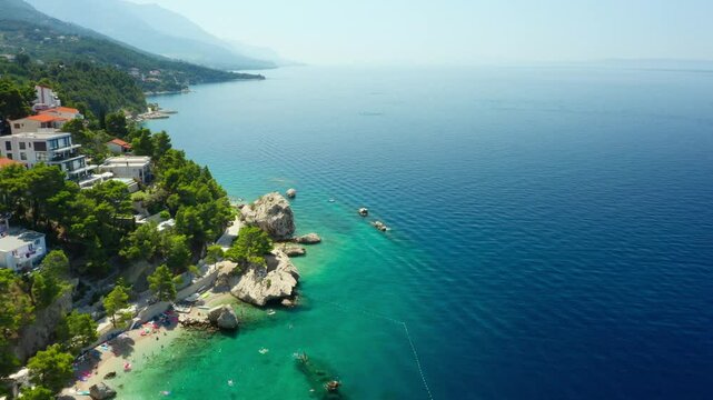Hotels and houses on the mountainside on the seafront. Tourists sunbathe on the beach, swim in the sea, and ride paddle boards. A seaside tourist town on the picturesque Adriatic coast of Croatia.