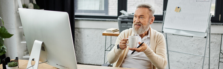 Mature handsome man sits at a desk, holding a coffee cup and looking at a computer screen.