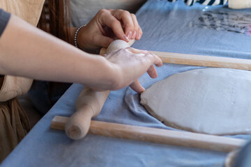 Close-up of a person's hands rolling out a slab of clay on a table during a pottery workshop. The scene captures the creative process with tools, rolling pins, and precision.