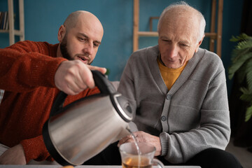 Family father and son drinking tea at home. Adult man pouring tea from teapot into cups for his elderly dad