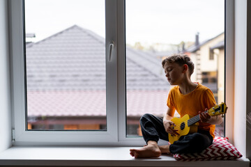 A boy dressed in an orange shirt focused on strumming a bright yellow ukulele in a cozy his room at...