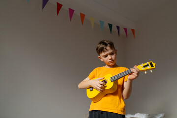 A boy dressed in an orange shirt focused on strumming a bright yellow ukulele in a cozy his room at...