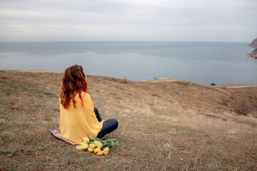 A woman sits on the grass with a bouquet of yellow flowers in her lap. She is looking out at the ocean, taking in the view. Concept of peace and tranquility.