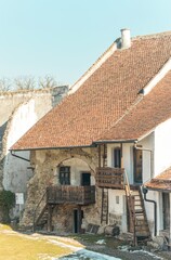 A quaint, rustic house showcases a blend of ancient stone and wooden architecture under a clear blue sky. The inviting balcony adds character, surrounded by serene greenery