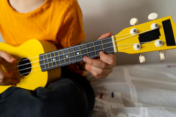 Obraz premium A boy dressed in an orange shirt focused on strumming a bright yellow ukulele in a cozy his room at home.