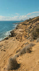 Rugged coastal terrain with rocky shoreline, calm sea, and distant clouds under a clear blue sky.