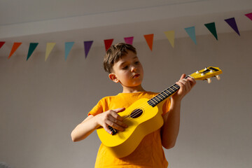 A boy dressed in an orange shirt focused on strumming a bright yellow ukulele in a cozy his room at...