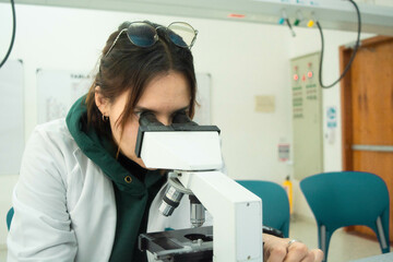 Young scientist woman in laboratory looking in a microscope, using lab coat. Lab background of...