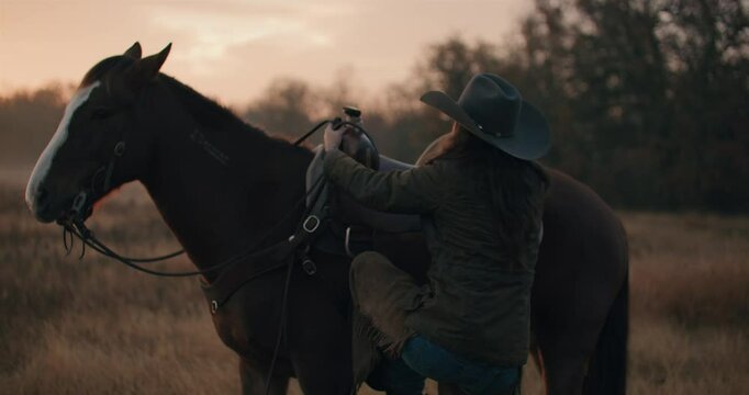 Woman Wearing Cowboy Hat Adjusting Saddle and Getting Onto Horse In Grassy Field At Golden Hour of Sunset