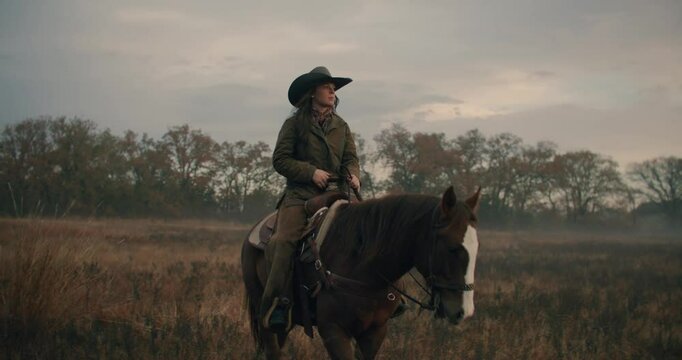 Cowgirl Woman Wearing Cowboy Hat and Holding Reins While Riding Horse and Walking Through Meadow With Sunset Cloudy Sky 