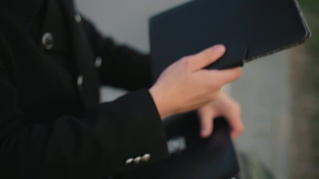 Close up of individual hand in black clothing opening briefcase, searching through documents, retrieves black book, background features interlocked path, stone structure, greenery in outdoor setting