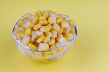 A transparent glass cup with ripe canned corn stands on a yellow background. Canned corn grains.