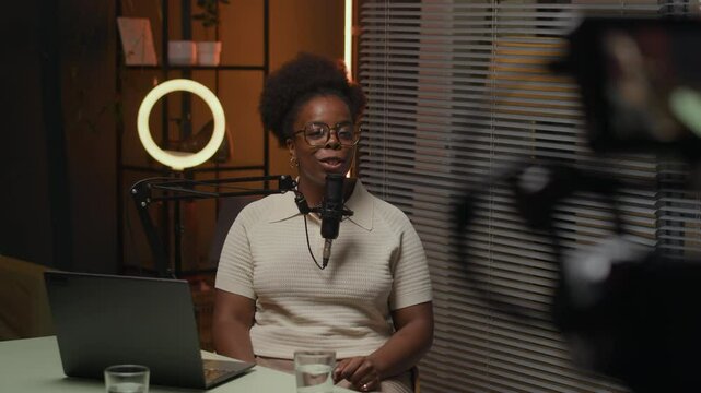 Backstage from podcasting with young smiling African American woman sitting at table in front of laptop and speaking to mic with laugh in cozy equipped broadcasting studio in front of filming camera