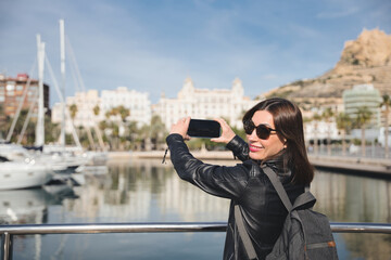 Smiling tourist taking pictures of the port of alicante with a smartphone, wearing sunglasses and a backpack, enjoying a sunny day in spain