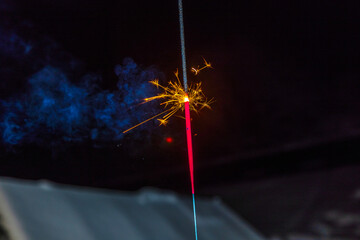 Burning sparkler emitting bright golden sparks with smoke trail against dark background at night. 