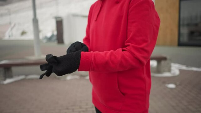 Woman putting on black winter glove for protection against cold outdoors, background shows modern benches and light pole, emphasizing winter urban landscape, she is dressed in a cozy red hoodie