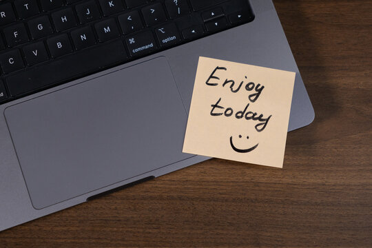 motivational sticky note with handwritten enjoy today message and smiley face on laptop trackpad, positive reminder