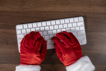santa claus typing on a white wireless keyboard in red gloves on a wooden desk background