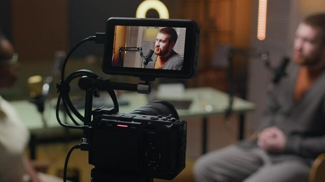 Cropped shot of cameraman adjusting exposure on camera set on tripod filming podcast with young Caucasian male guest speaking in mic and Black female host in blurred background in cozy studio