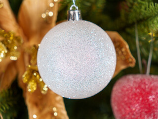 A reflective white ornament adorned with glitter hangs delicately on a Christmas tree. Surrounding...