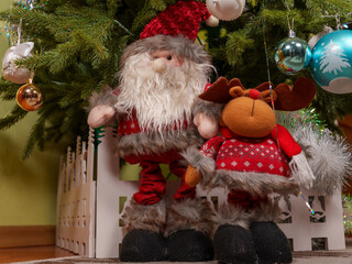 Colorful ornaments hang from a Christmas tree, beneath which stand plush Santa and reindeer figures...
