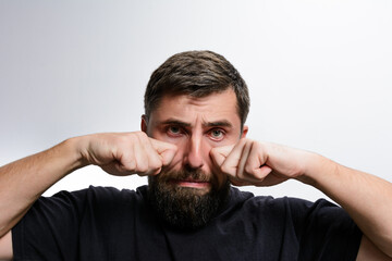 A man with a beard rubs his eyes with his fists against a light gray background. He appears tired, possibly upset, or experiencing eye discomfort.