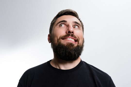 A man with a beard looks upwards with a curious and slightly mischievous expression on his face against a plain background.