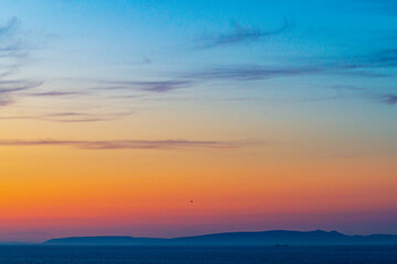 Beautiful sunset landscape of ocean or sea, stretching into distance to other shore with hills. Blurred photo due to haze rising from surface of water. Contrast of blue and orange colors in nature