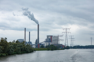 Obraz premium Industrial power plant with smoke stacks and transmission towers by a river. Gray overcast sky.