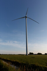 Fototapeta premium Large wind turbine dominates a Dutch landscape, blue sky, and grassy fields. Renewable energy in rural setting.