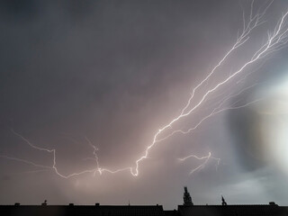 Dramatic lightning illuminates a gray sky above rooftops.  Powerful storm activity.