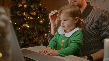 Cute little child sitting with her father and working on the computer with a Christmas tree in the background.