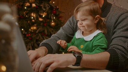 Cute little child sitting with her father and working on the computer with a Christmas tree in the background.