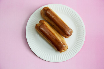 Two éclairs are lying on a white paper plate against a pink background in close-up, a delicate dessert, sweets and treats, cream-filled pastry.
