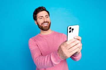 Cheerful bearded man in pink sweater taking a selfie with smartphone against blue background, showcasing modern lifestyle and technology