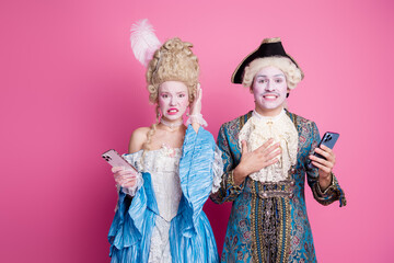Two people dressed in elegant historical costumes against a pink background, holding smartphones and expressing surprise.