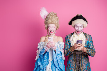 Elegant couple in historical costumes enjoying smartphone entertainment against a vibrant pink background at a theme party